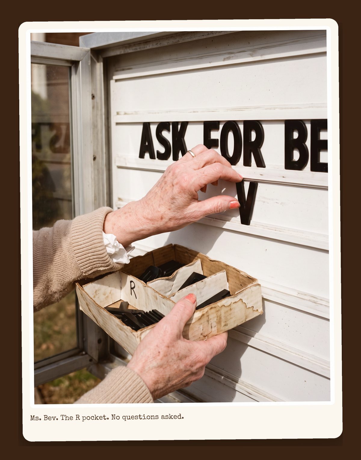 Ms. Bev's hands at the sign's letter box, sliding a black plastic letter into a white track, the R pocket of the letter box empty.