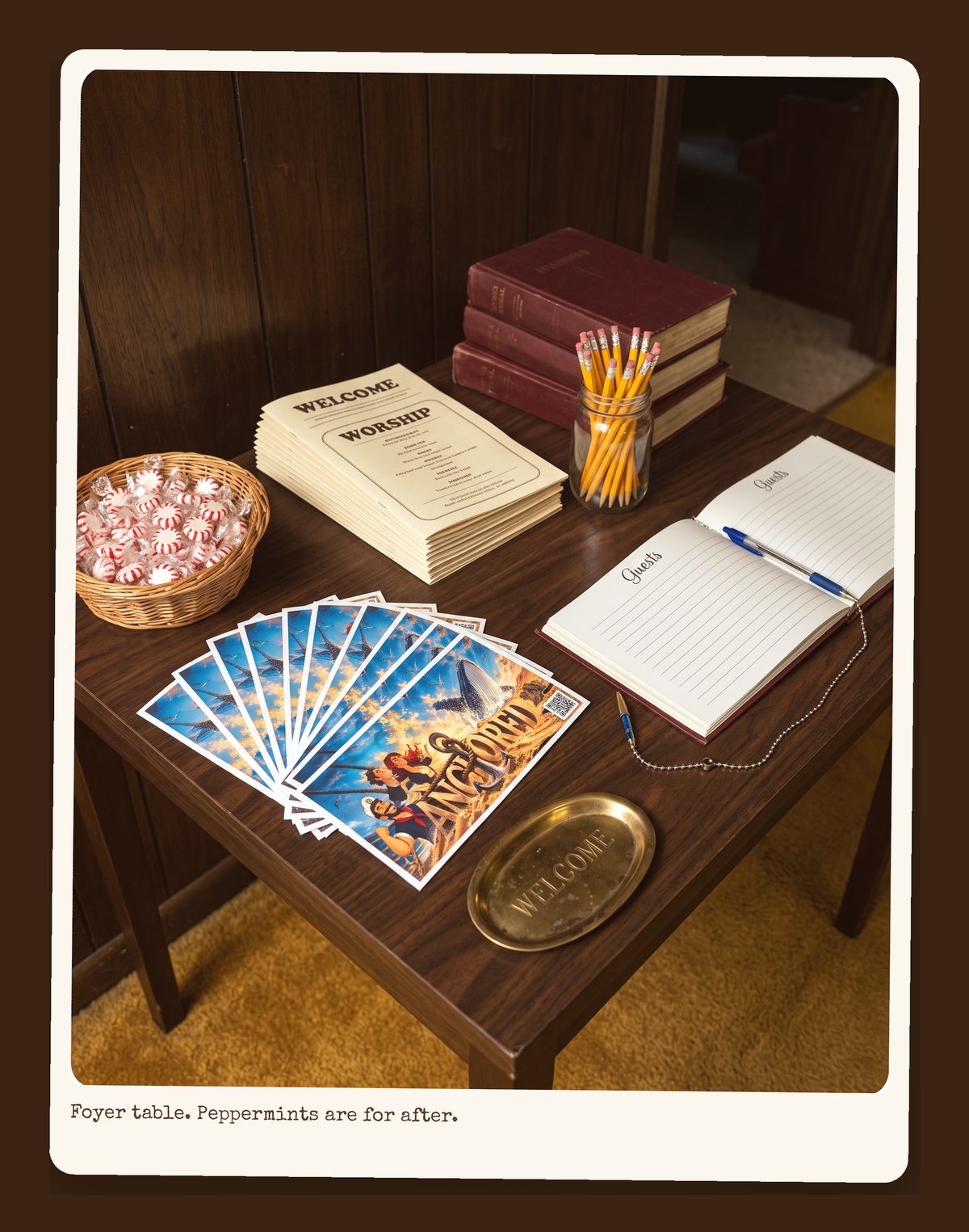 The welcome table: a stack of bulletins, hymnals, a bowl of peppermints, a sign-in sheet with a pen on a chain, and a fan of Anchored bulletin inserts.